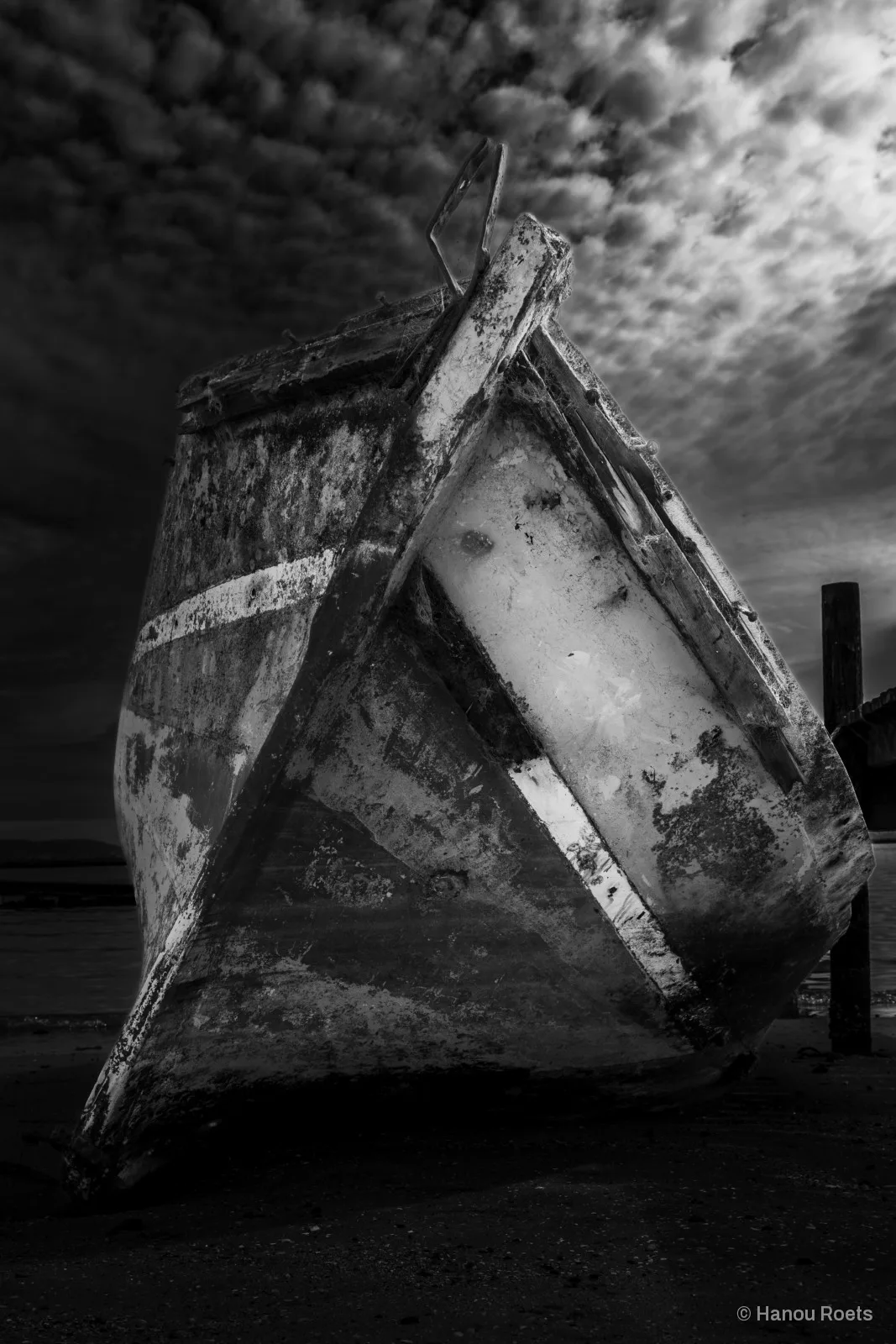 The peeling, weathered bow of an old wooden boat resting on dark sand beneath a dramatic mottled sky, with a wooden post to one side