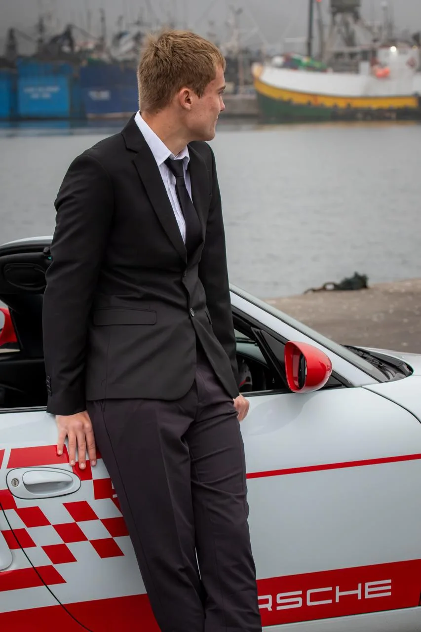 A young man in a suit leaning on a white and red Porsche at the harbour