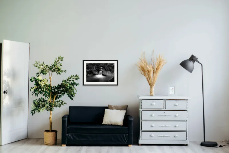 Forest falls shown framed as an A2 print, above a black two-seater between a potted tree and a chest of drawers