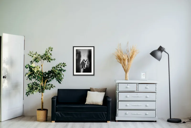 Three’s company shown framed as an A2 print, above a black two-seater between a potted tree and a chest of drawers
